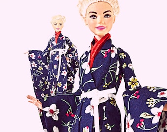 May include: A doll with blonde hair wearing a dark blue kimono featuring a white and red floral pattern. The garment includes a red inner collar and a white waist tie. The doll has a light skin tone, painted facial features, and wide sleeves.