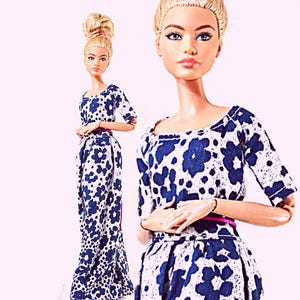 Puede incluir: Una muñeca con cabello rubio recogido en un moño alto, que viste un vestido de manga larga con un estampado floral azul oscuro sobre fondo blanco. El vestido tiene un escote redondo y un cinturón fino de color rosa en la cintura. La muñeca tiene brazos articulados.