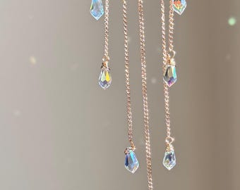 Puede incluir: Cadenas doradas con colgantes de cristal iridiscente cuelgan sobre un fondo neutro. Los cristales, con forma de lágrima, capturan la luz, creando un efecto brillante. Las finas cadenas añaden elegancia a la pieza, creando una apariencia etérea.