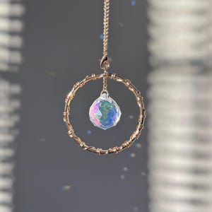 Puede incluir: Un atrapasoles de metal dorado con una bola de cristal transparente y facetada suspendida dentro de un marco circular. El marco está envuelto con alambre, y el atrapasoles cuelga de una delicada cadena. La bola de cristal refleja un arcoíris de colores.