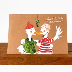 Könnte beinhalten: Eine Weihnachtskarte mit zwei Cartoon-Gänsen, die Hüte und Pullover tragen und sich unter einem Zweig mit Stechpalme küssen. Die Karte ist braun und hat den Text "ganz friedliche Weihnachten" in weiß.
