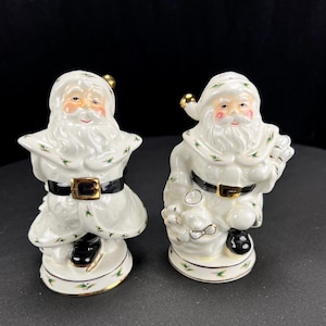 May include: Two ceramic Santa Claus figurines. Each Santa is wearing a white suit with a black belt and gold buckle. The figurines have rosy cheeks, white beards, and green and gold accents. One Santa is holding a sack.