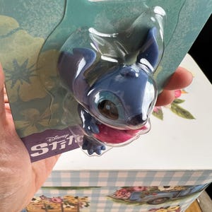 May include: A blue Stitch figurine in a clear plastic package. The figurine has large ears, a pink tongue, and is from Disney's Stitch franchise. The packaging has the Disney Stitch logo.
