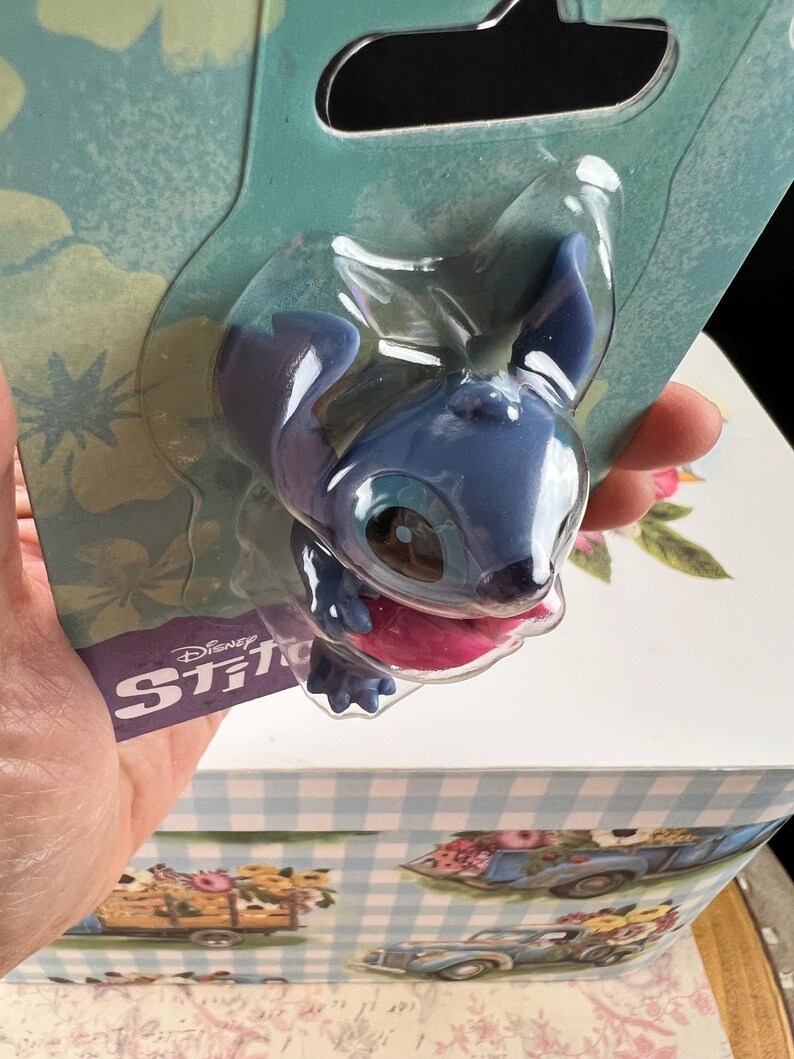 May include: A blue Stitch figurine in a clear plastic package. The figurine has large ears, a pink tongue, and is from Disney's Stitch franchise. The packaging has the Disney Stitch logo.