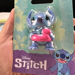 May include: A blue Stitch figurine with a pink inflatable ring, packaged on a teal and purple card. The card features floral patterns and the Disney Stitch logo. The packaging indicates the toy is suitable for ages 3+.