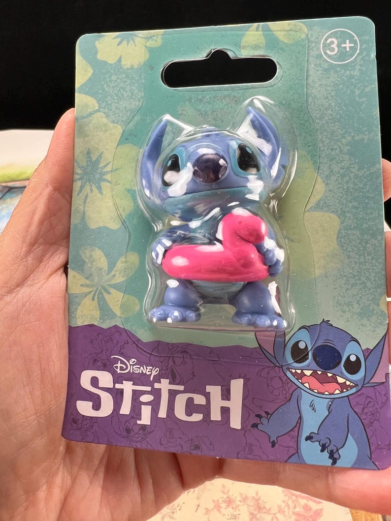 May include: A blue Stitch figurine with a pink inflatable ring, packaged on a teal and purple card. The card features floral patterns and the Disney Stitch logo. The packaging indicates the toy is suitable for ages 3+.