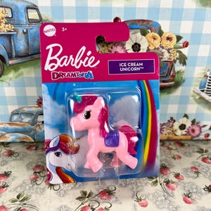May include: A Barbie Dreamtopia Ice Cream Unicorn toy in a clear plastic package. The pink unicorn has a purple saddle and a rainbow mane and tail. The packaging is pink with the Barbie logo and the text "Ice Cream Unicorn".
