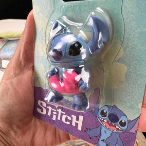 May include: A blue Stitch figurine in a clear plastic package. The figurine has large ears, a pink object, and the word "Stitch" in white on a purple background. A cartoon image of Stitch is also on the packaging.