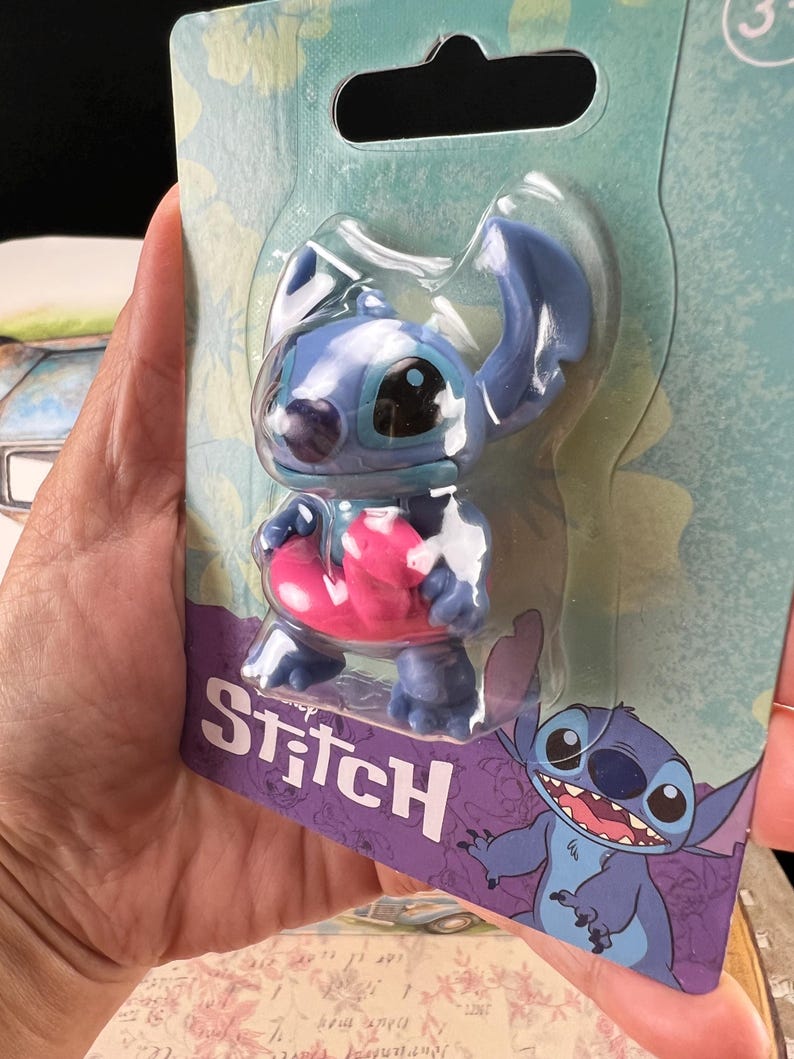 May include: A blue Stitch figurine in a clear plastic package. The figurine has large ears, a pink object, and the word "Stitch" in white on a purple background. A cartoon image of Stitch is also on the packaging.