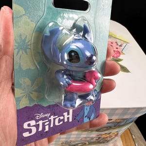 May include: A blue Stitch figure with a pink inflatable ring, presented in a clear plastic blister pack. The packaging displays the Disney Stitch logo and a blue and purple floral pattern.