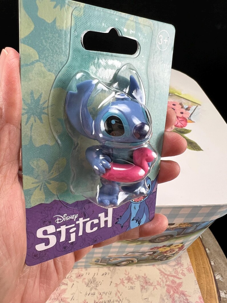May include: A blue Stitch figure with a pink inflatable ring, presented in a clear plastic blister pack. The packaging displays the Disney Stitch logo and a blue and purple floral pattern.