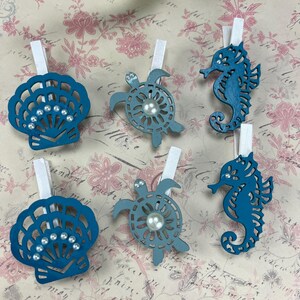 May include: A set of six teal wooden clothespins featuring cutout shapes of two seashells, two sea turtles, and two seahorses. Each shape is decorated with small white faux pearls. The clothespins are arranged in two rows of three on a patterned surface.