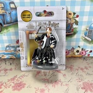 May include: A Harry Potter figurine in a clear plastic package. The figurine depicts a character in a black robe, holding a wand. The packaging includes the text "Harry Potter" and "Bonus Card!"