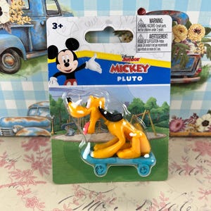 Puede incluir: Una figura de Pluto sobre una patineta azul, empaquetada en un blíster transparente. El embalaje presenta a Mickey Mouse y las palabras "Mickey Junior Pluto". El fondo es un estampado floral y de cuadros azules.