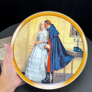 May include: A decorative plate featuring a romantic scene with a man in a blue cape whispering to a woman in a white dress. The plate has a gold rim and is signed "Norman Rockwell".