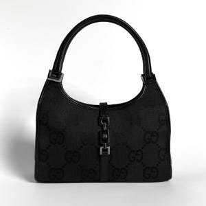 May include: A black handbag featuring a tonal GG monogram pattern across the fabric body. The bag includes a single curved top handle, black leather trim, and a central silver-toned metal piston-style closure mechanism on the front panel.