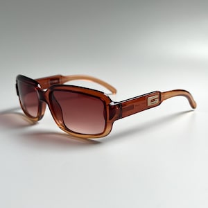 Puede incluir: Gafas de sol marrones con montura rectangular y lentes degradadas. Las gafas de sol tienen un logotipo dorado en las patillas. Las patillas son de color marrón claro. Diseñadas para proteger los ojos del sol.