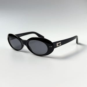May include: Black oval-shaped sunglasses with dark grey lenses. The frames feature a glossy finish and a silver-toned metal G logo on each temple. Two small silver rivets are visible on the outer corners of the frame front near the hinges.