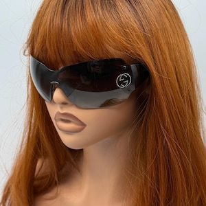 May include: Wraparound shield sunglasses featuring a dark gradient lens and a black frame. The left side of the lens displays a circular logo composed of small, clear crystals forming the interlocking double G symbol of the Gucci brand.