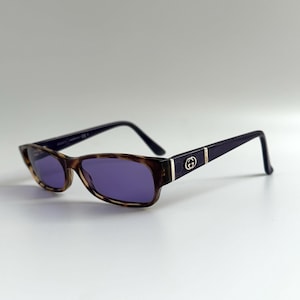 May include: Rectangular sunglasses featuring a brown tortoiseshell frame front and dark purple lenses. The temples are dark purple with a silver-toned metal Gucci logo emblem and a subtle repeating pattern embossed on the side near the hinges.