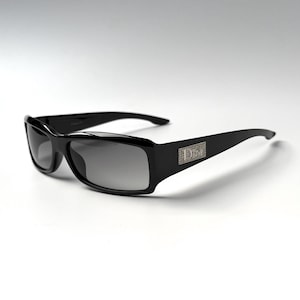 May include: Black sunglasses with rectangular frames and dark gradient lenses. The temples feature a rectangular silver-toned metal plaque embedded with small clear crystals that form the word Dior in a stylized font on the side of the frame.