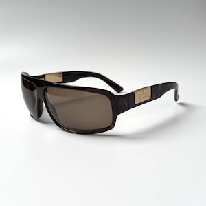 May include: Tortoiseshell-patterned sunglasses with a wrap-around frame and dark brown lenses. The temples feature a gold-tone metal plaque engraved with the text GUCCI MADE IN ITALY. The design includes a thick bridge and curved temple arms.