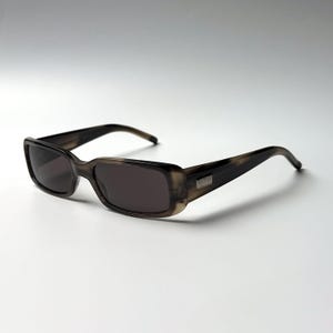 May include: Rectangular sunglasses with a dark grey and brown tortoiseshell pattern frame. The lenses are dark grey and tinted. A small silver-toned metal plaque with the Gucci logo is embedded on the outer temple of the frame near the hinge.