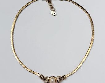 May include: A gold-tone chain necklace featuring a flat, braided-style link design. The center focal point is an oval-shaped faux pearl surrounded by small clear crystals. The necklace includes an adjustable chain with a CD logo charm at the end.