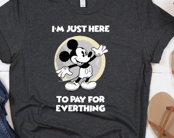 May include: Dark gray t-shirt featuring a Mickey Mouse graphic inside a yellow circle, with the text "I'M JUST HERE TO PAY FOR EVERYTHING." The shirt is paired with blue jeans and brown sandals, suggesting casual wear.