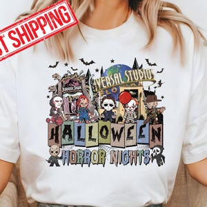 May include: A white t-shirt with a graphic print of a Halloween scene. The scene features a group of horror movie characters in front of a Universal Studios Halloween Horror Nights sign. The characters include Freddy Krueger, Jason Voorhees, Michael Myers, Pennywise, and Chucky. The text on the sign reads 'Universal Studios Halloween Horror Nights'. The t-shirt is being worn by a person with blue jeans.