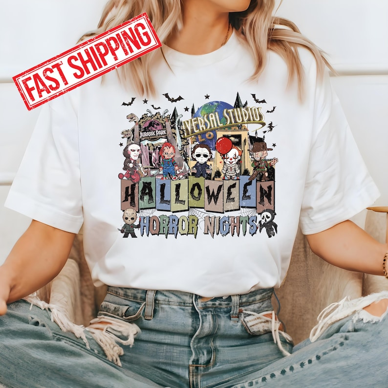 May include: A white t-shirt with a graphic print of a Halloween scene. The scene features a group of horror movie characters in front of a Universal Studios Halloween Horror Nights sign. The characters include Freddy Krueger, Jason Voorhees, Michael Myers, Pennywise, and Chucky. The text on the sign reads 'Universal Studios Halloween Horror Nights'. The t-shirt is being worn by a person with blue jeans.