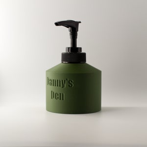 May include: An olive green soap dispenser with a black pump and collar. The words "Danny's Den" are embossed on the front of the dispenser. The dispenser is cylindrical with a slightly conical top.