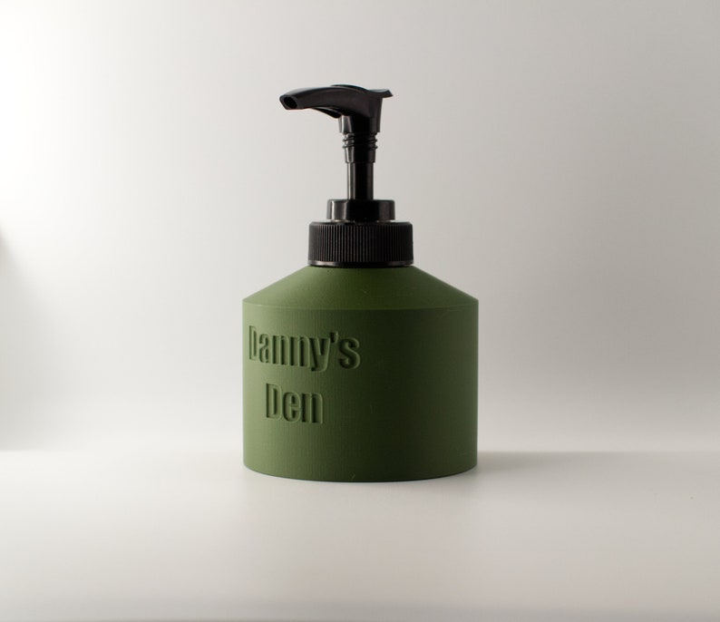 May include: An olive green soap dispenser with a black pump and collar. The words "Danny's Den" are embossed on the front of the dispenser. The dispenser is cylindrical with a slightly conical top.