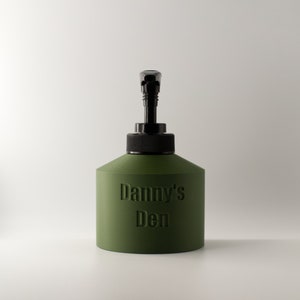 May include: An olive green dispenser bottle with a black pump and cap. The embossed text "Danny's Den" is visible on the front. The bottle has a rounded, geometric shape and is set against a white background.