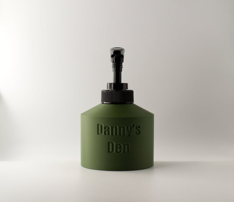 May include: An olive green dispenser bottle with a black pump and cap. The embossed text "Danny's Den" is visible on the front. The bottle has a rounded, geometric shape and is set against a white background.