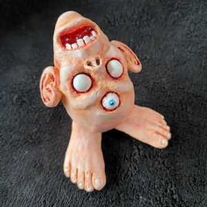 Puede incluir: Una escultura grotesca, hecha a mano, de una figura pálida y carnosa con múltiples ojos, una boca muy abierta con encías rojas y dientes blancos, y dos pies. La figura tiene una apariencia perturbadora.