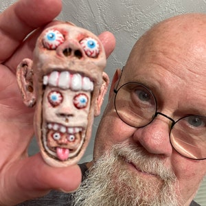 May include: A hand holds a small, ceramic figurine of a grotesque face with multiple eyes and a wide, toothy grin. The figurine is painted in shades of pink, white, and blue.
