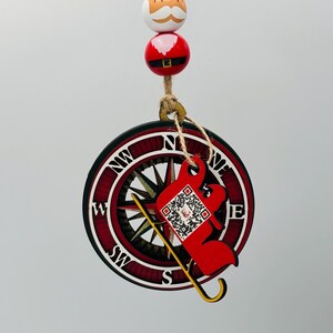 May include: Red and white wooden Christmas ornament featuring a compass with a Santa Claus in a sleigh with a QR code. The ornament is hanging from a string with a Santa Claus bead.