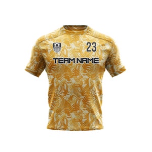 May include: A gold soccer jersey with a fern leaf pattern. The jersey has a black team logo, the number 23, and the text "TEAM NAME".