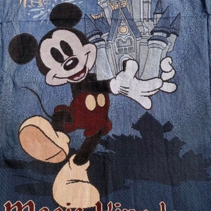 Puede incluir: Un tapiz azul y blanco con Mickey Mouse de pie frente al Castillo de Cenicienta con fuegos artificiales en el fondo. El texto "Magic Kingdom" est&aacute; en la parte inferior del tapiz.