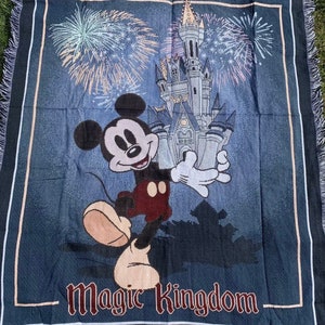 Puede incluir: Una manta tejida azul con un personaje de Mickey Mouse de pie frente al Castillo de Cenicienta. El castillo est&aacute; rodeado de fuegos artificiales y el texto "Magic Kingdom" est&aacute; en la parte inferior de la manta. La manta tiene un borde de flecos.