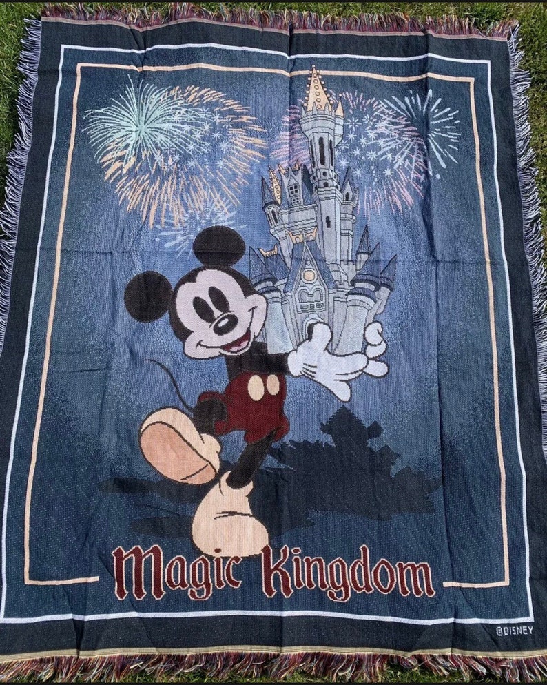 Puede incluir: Una manta tejida azul con un personaje de Mickey Mouse de pie frente al Castillo de Cenicienta. El castillo est&aacute; rodeado de fuegos artificiales y el texto "Magic Kingdom" est&aacute; en la parte inferior de la manta. La manta tiene un borde de flecos.