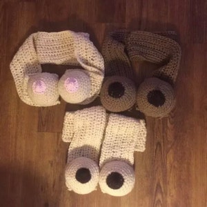 Puede incluir: Artículos de novedad hechos a mano con ganchillo, que incluyen una bufanda beige con dos accesorios en forma de pecho, una bufanda marrón con dos accesorios en forma de ojo y un par de mitones blancos con accesorios en forma de ojo. Los artículos están hechos de hilo y están colocados sobre una superficie de madera.