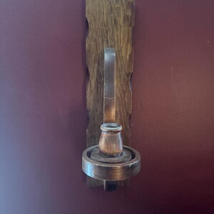 Puede incluir: Un candelabro de pared de madera que presenta un tablero rectangular con bordes festoneados, un brazo de soporte curvo y una base circular con una pequeña copa central para sostener una vela. Toda la pieza tiene un acabado de tinte marrón oscuro.