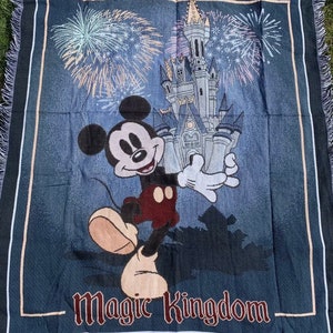 Puede incluir: Una manta de tejido azul con un personaje de Mickey Mouse de pie frente al Castillo de Cenicienta. El castillo est&aacute; rodeado de fuegos artificiales y el texto "Magic Kingdom" est&aacute; impreso en la parte inferior de la manta. La manta tiene un borde de flecos.