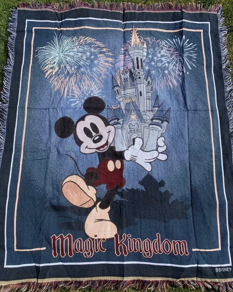 Puede incluir: Una manta de tejido azul con un personaje de Mickey Mouse de pie frente al Castillo de Cenicienta. El castillo est&aacute; rodeado de fuegos artificiales y el texto "Magic Kingdom" est&aacute; impreso en la parte inferior de la manta. La manta tiene un borde de flecos.