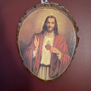 Puede incluir: Una placa de pared con la imagen impresa de una figura con cabello largo castaño y barba, vistiendo una túnica blanca y una túnica roja. La figura sostiene un corazón rojo con una cruz encima. La impresión está montada sobre un rodaja de madera.