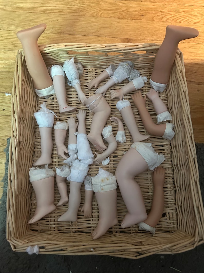Puede incluir: Una colecci&oacute;n de extremidades de mu&ntilde;ecas de porcelana, incluyendo piernas y brazos, se encuentra dentro de una cesta de mimbre tejida. Las extremidades var&iacute;an en tono de piel y est&aacute;n parcialmente vestidas con tela y encaje.