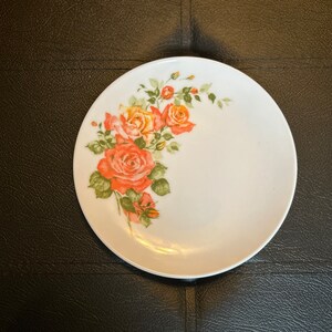May include: A white ceramic plate with a floral design featuring orange and yellow roses with green leaves. The roses are clustered on the left side of the plate.