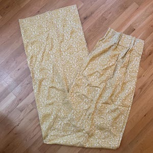 Puede incluir: Un panel de cortina largo y rectangular con un patrón de damasco repetitivo en color amarillo mostaza y blanco. La tela muestra un diseño floral intrincado y ornamentado en toda su superficie, con un encabezado fruncido y plisado en el borde superior.